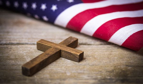 A holy wooden Christian cross laying on a wood background with an American flag. File photo credit: enterlinedesign via Shutterstock.