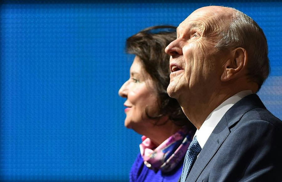 An elderly man in a suit and tie looks up, positioned against a blue background, as a brunette woman with a pink scarf sits beside him.