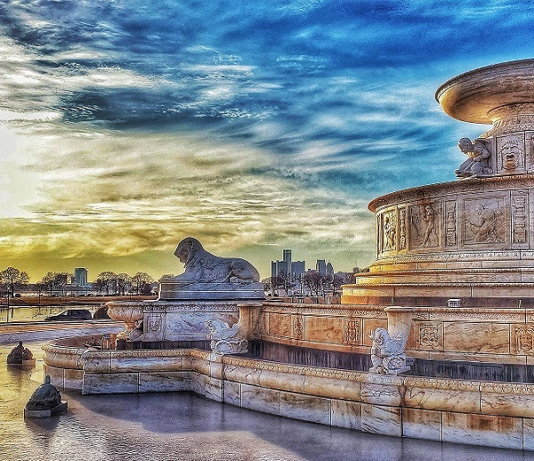 brilliant sunrise lights up a bright blue, cloudy sky behind an ornate concrete, circular fountain with carved lions and tiers