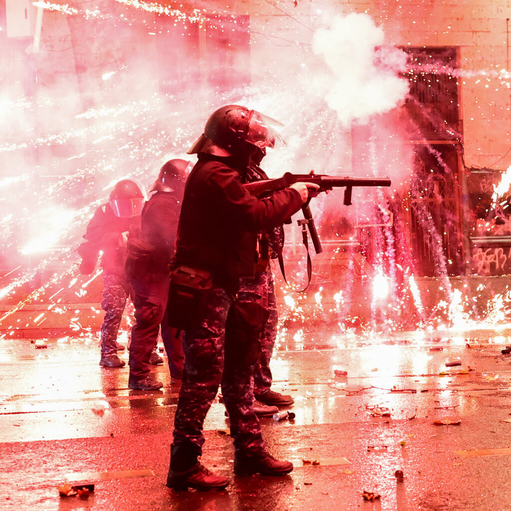 Police officers in riot gear amid explosions. 