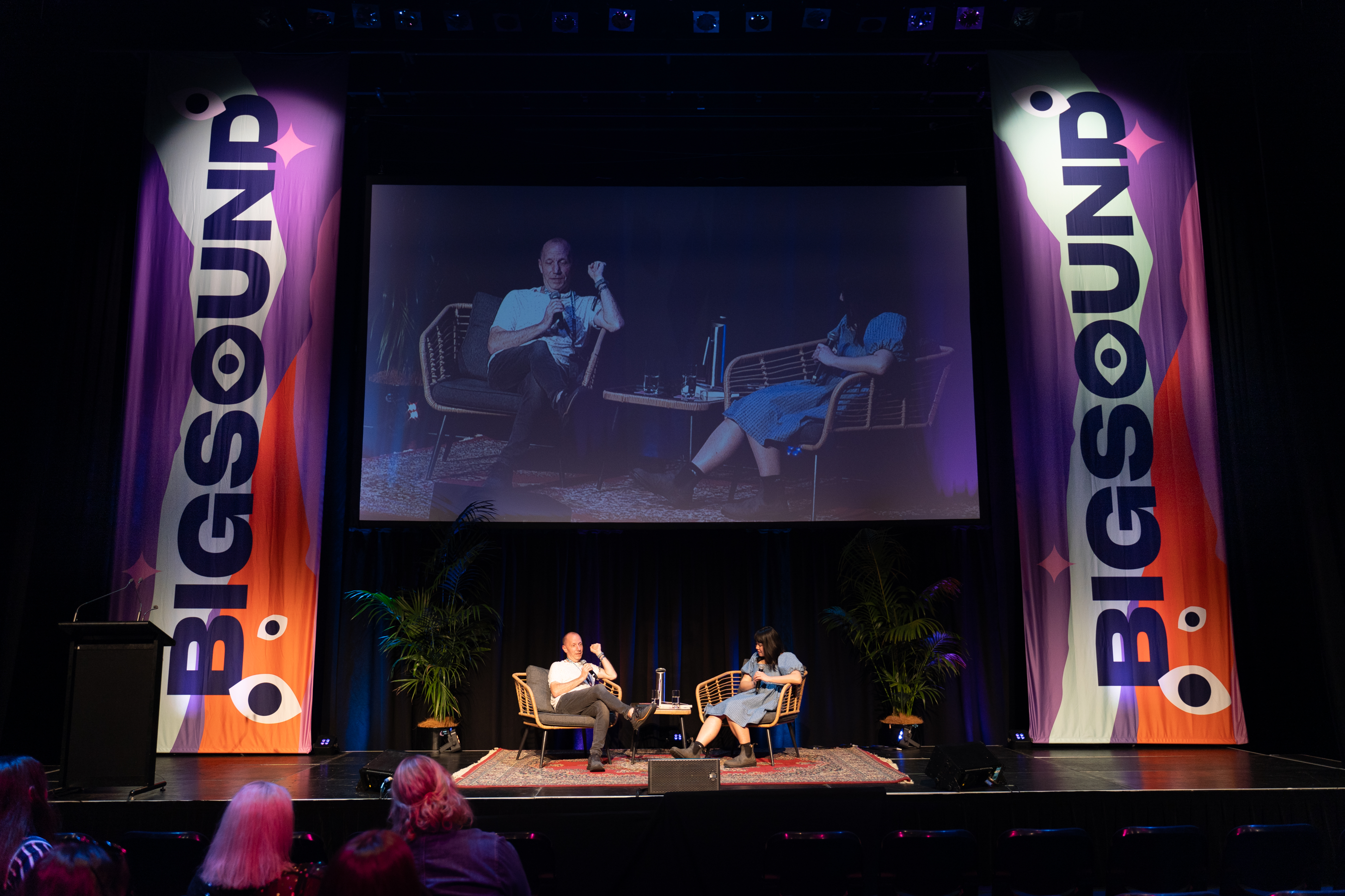 A man and woman sitting on chairs on stage underneath a large screen. On both sides of the stage are large banners with the BIGSOUND logo.
