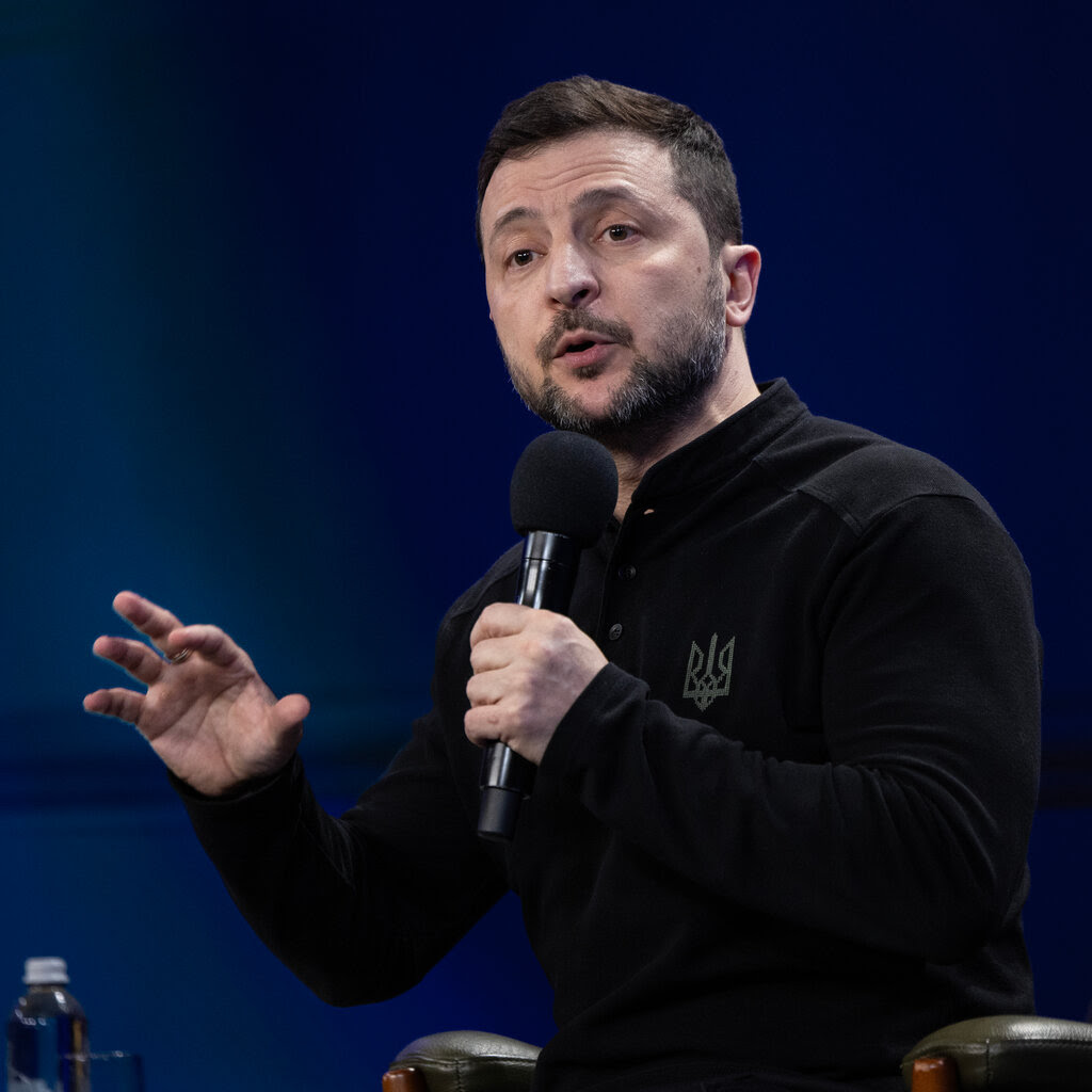 Volodymyr Zelensky in a black shirt speaking into a microphone.