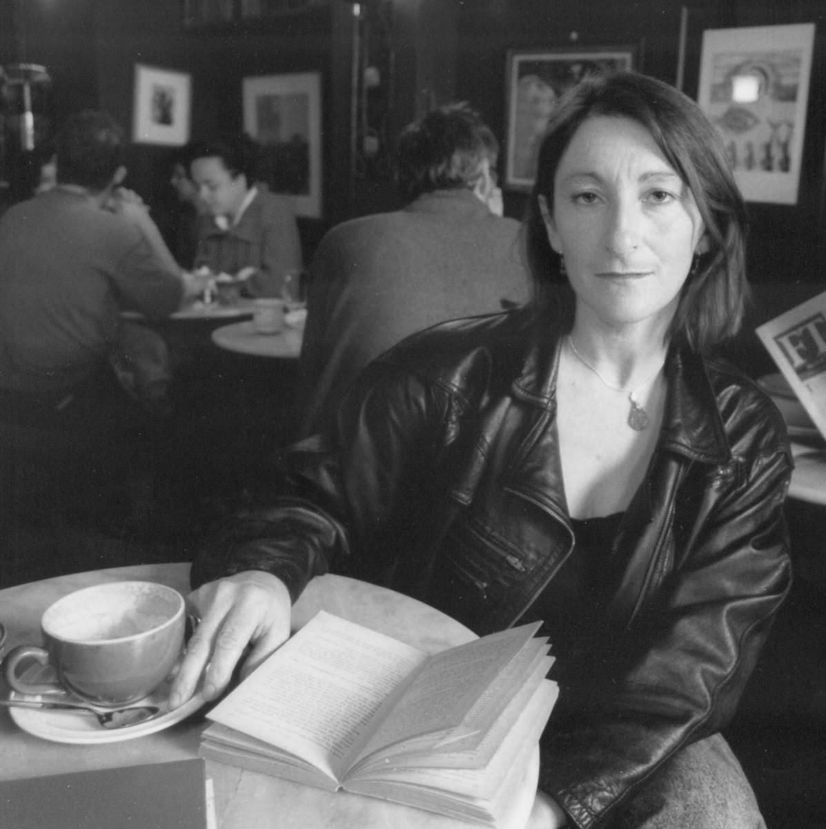 In this black-and-white image, poet Mary O'Malley sits at a table in a cafe, an open book before her. She is staring intently at the camera.