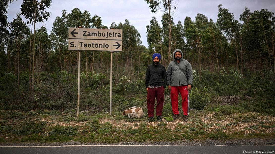 Dois homens posam à beira de estrada ao lado de placas indicando cidades Zambujeira e S. Teotónio, com bosque ao fundo