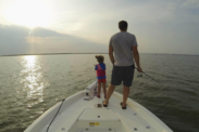 Parent holding a fishing pole and a child in a life jacket standing on the bow of a fishing boat at sunset on calm water.