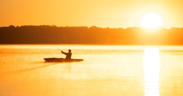A fisherman, silhouetted in the glow of an early sunrise, casts a line from a kayak on a placid, misty lake.