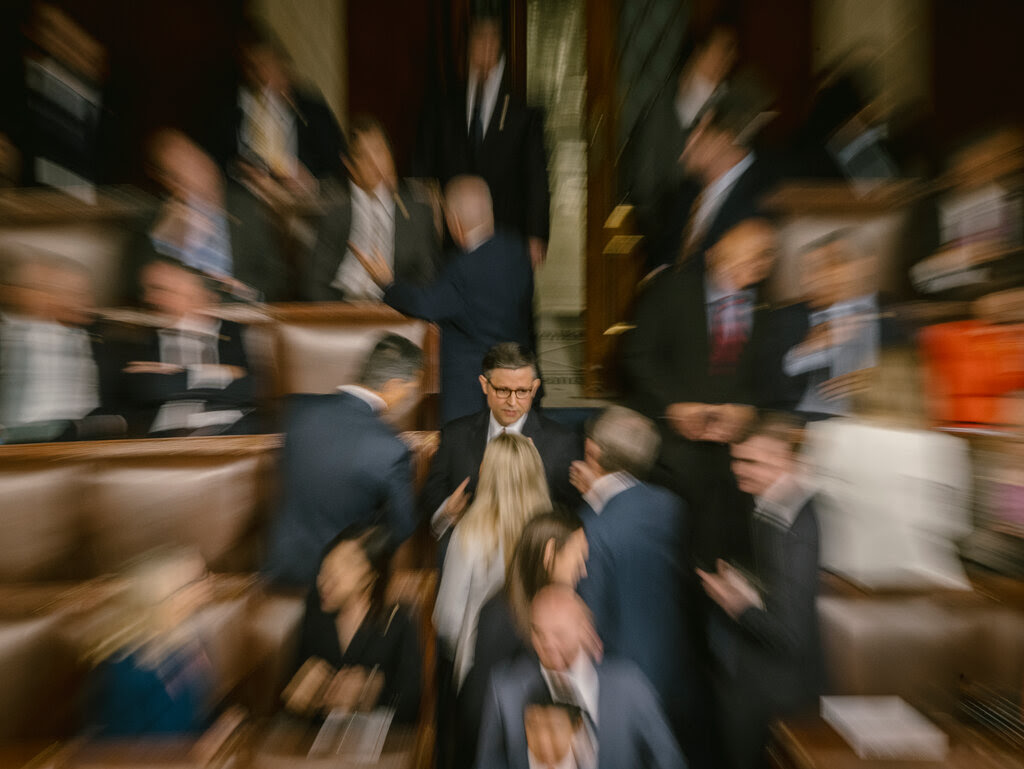 Speaker Mike Johnson speaks with other people in an aisle between rows of seats at the U.S. Capitol. The photograph is focused on him at the center, while those around him appear blurred.