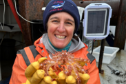 Dr. Alix LaFerriere wears warm fishing gear aboard a boat. She is smiling at the camera holding up a crab