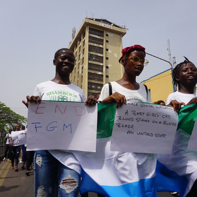 Three young women in white T-shirts stand on an urban street, holding homemade signs with messages including “End FGM” (for female genital mutilation).