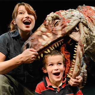 A puppet dinosaur holds its jaws around a smiling child's head.