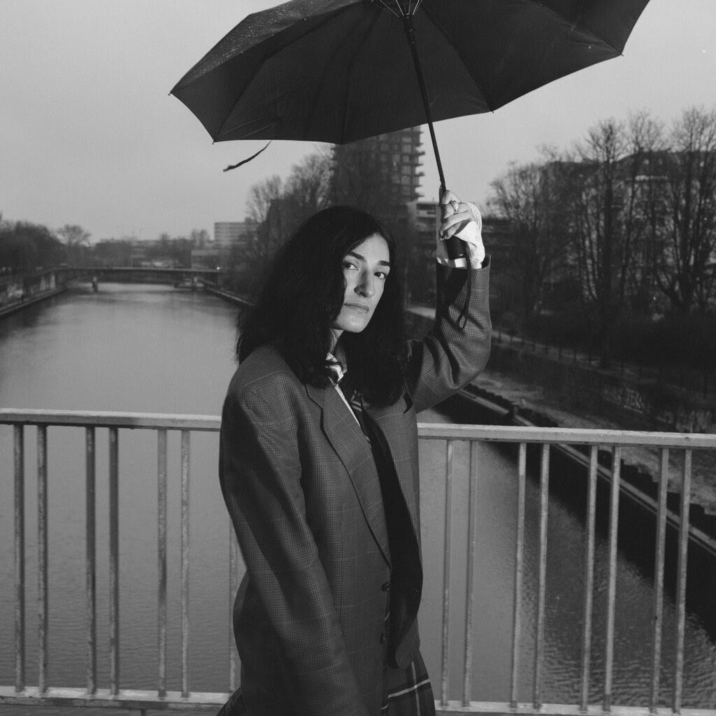 A black-and-white photograph of Moshtari Hilal, who is standing on a bridge over a waterway and holding an umbrella over her head. 