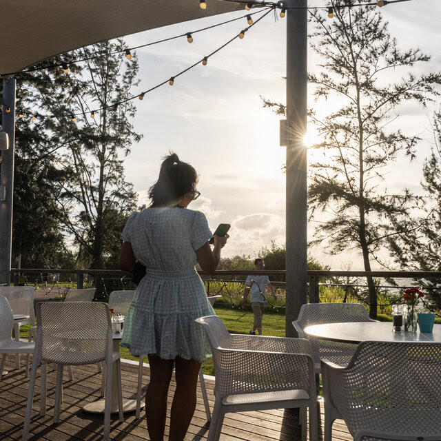 A figure in a short dress and a high ponytail looks at her phone standing among some cafe chairs.