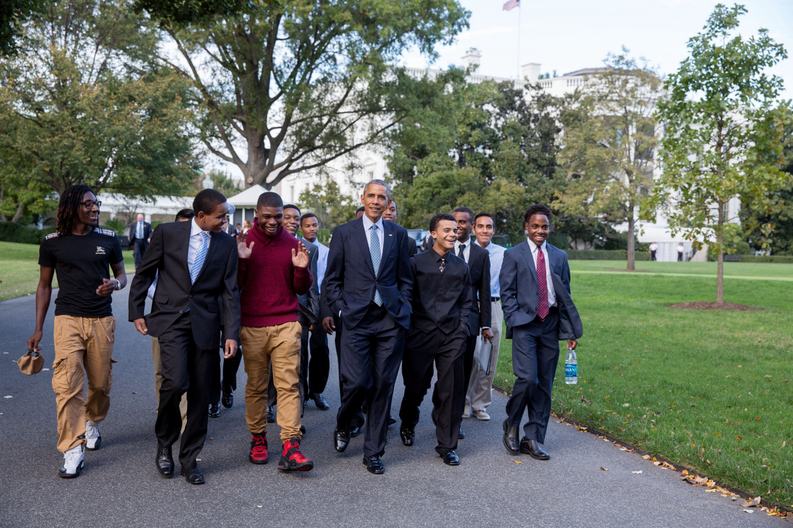 President Obama is wearing a suit and walking on a path in front of the White House alongside a group of Black young men with a range of skintones and hairstyles.