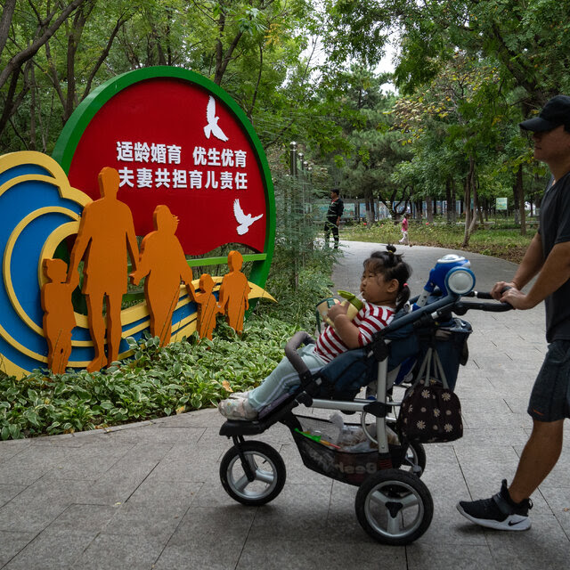 In a park, a family walks past artwork featuring life-size cutout of a man and woman walking with three children, under a slogan urging couples not to wait too long to have children.