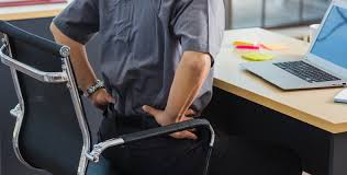 A man sitting uncomfortably in his office chair, grimacing slightly as he tries to adjust his posture. He's leaning forward awkwardly, his hands on his lower back, indicating discomfort