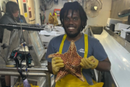 Jonathan wearing a grey t-shirt and yellow coveralls while holding a big red cushion sea star