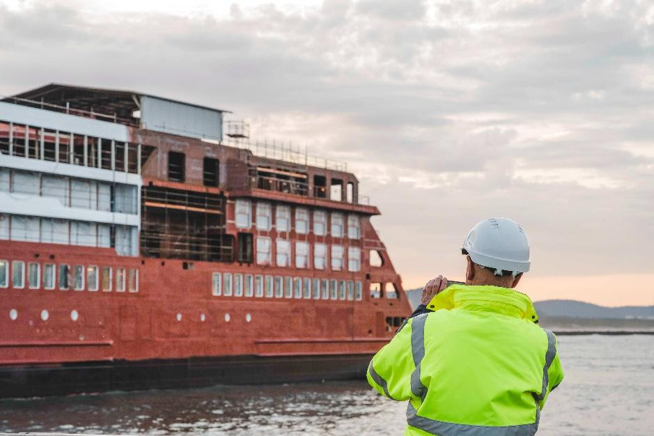 A person in a yellow vest and a white helmet standing in front of a large shipDescription automatically generated