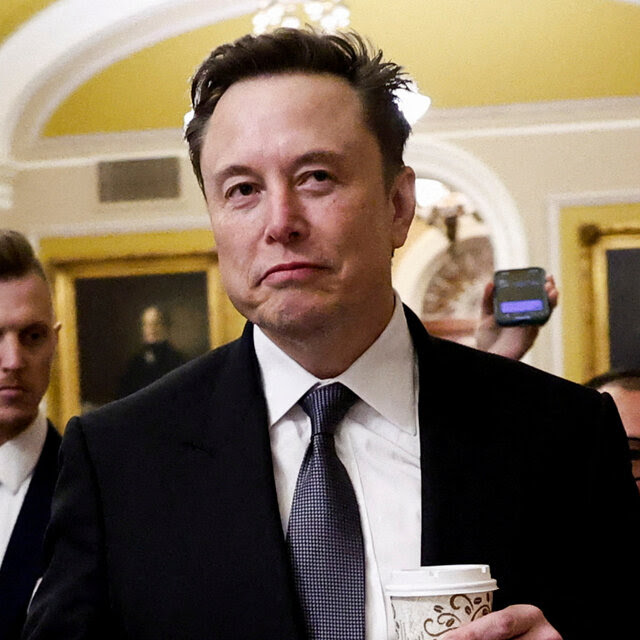 Elon Musk in a dark suit holding a paper takeaway cup. 