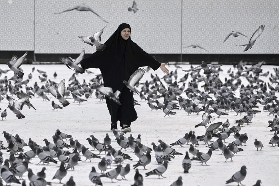 A woman wearing black, flowing clothes and a hijab walks through a flock of pigeons.