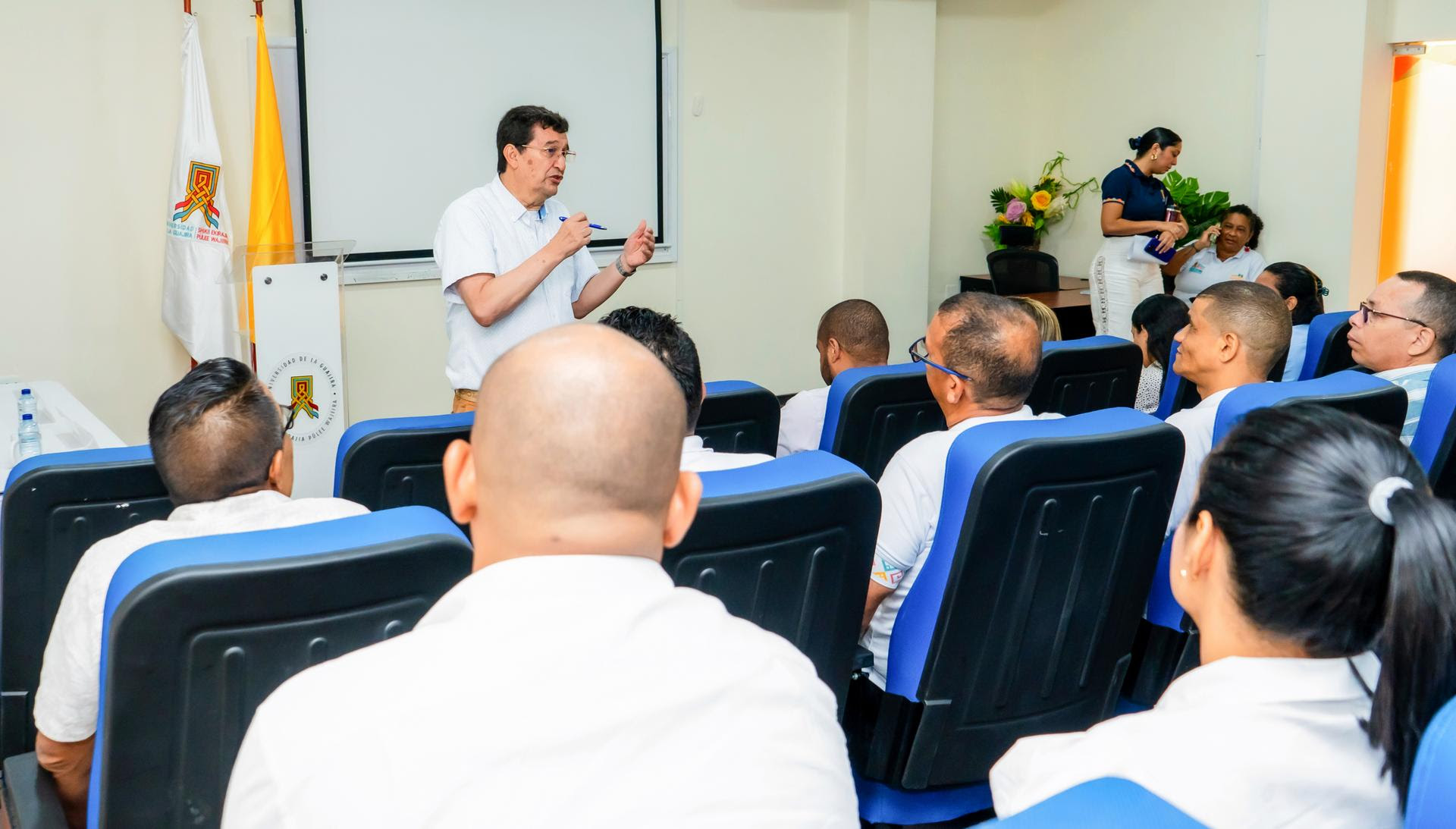 Un conferencista imparte una charla a un grupo de personas sentadas en una sala de conferencias, con las banderas institucionales de fondo.