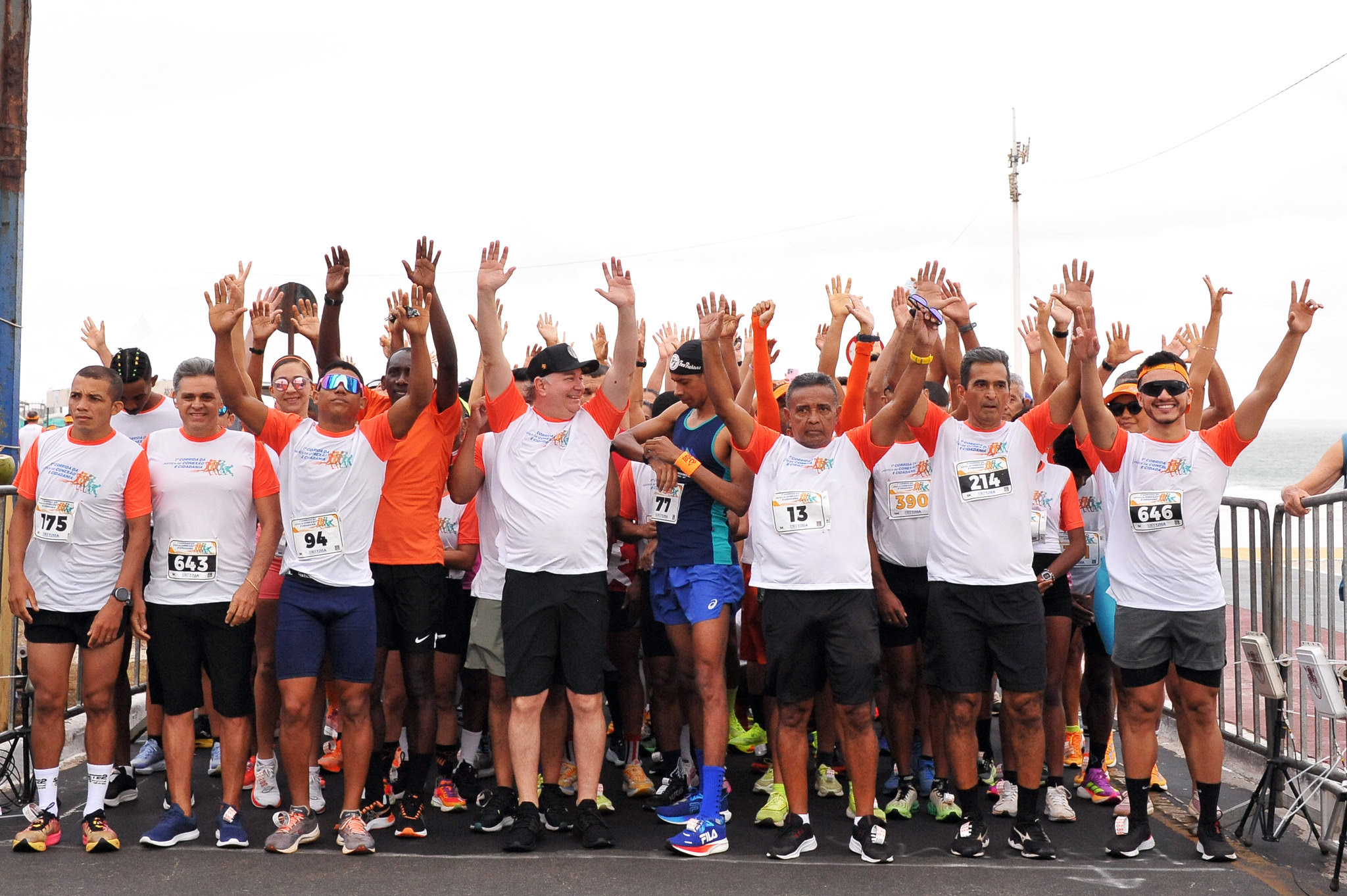 A imagem mostra um grupo de pessoas em um evento esportivo, uma corrida, sugerido pela presença de números de identificação em alguns participantes. As pessoas estão levantando as mãos, o que pode indicar celebração ou participação em uma atividade.