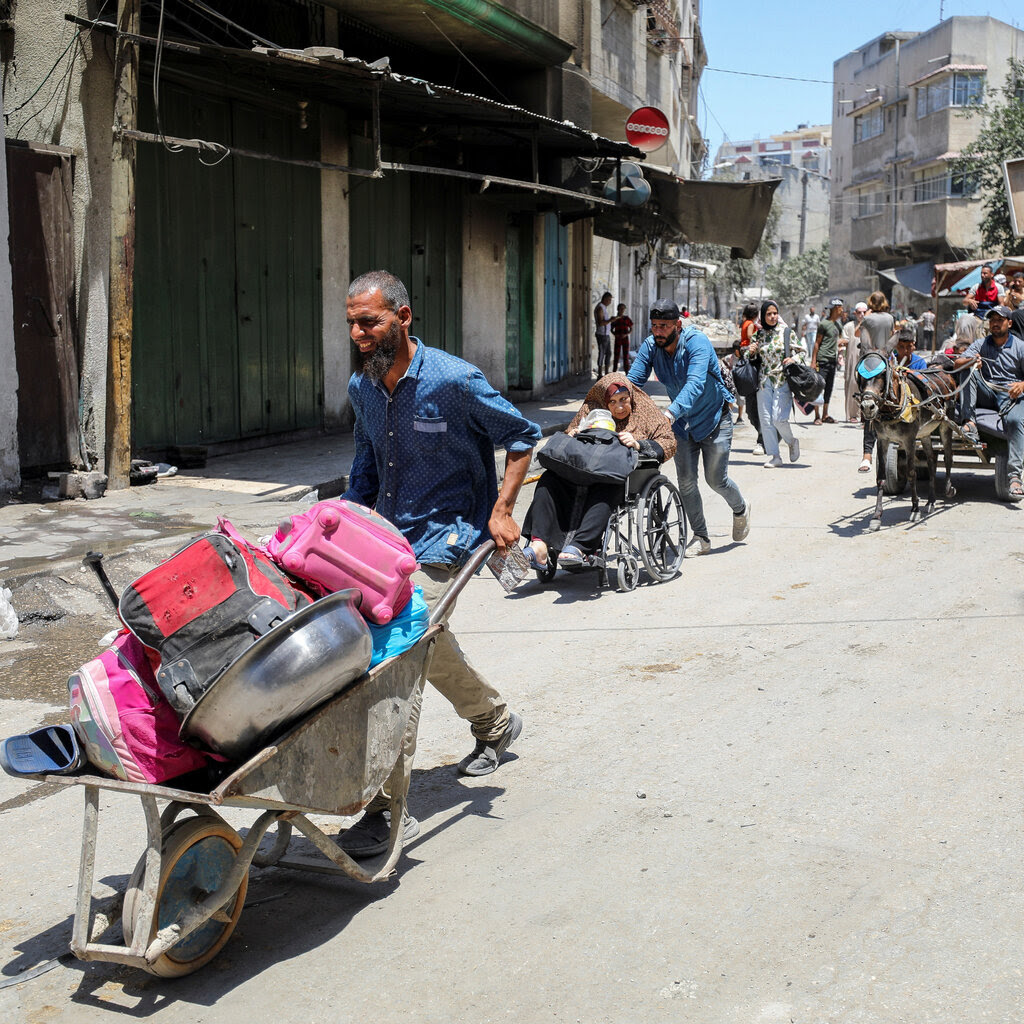 A Palestinian man with a wheel barrel in the streets with a man pushing a woman in a wheelchair nearby. 