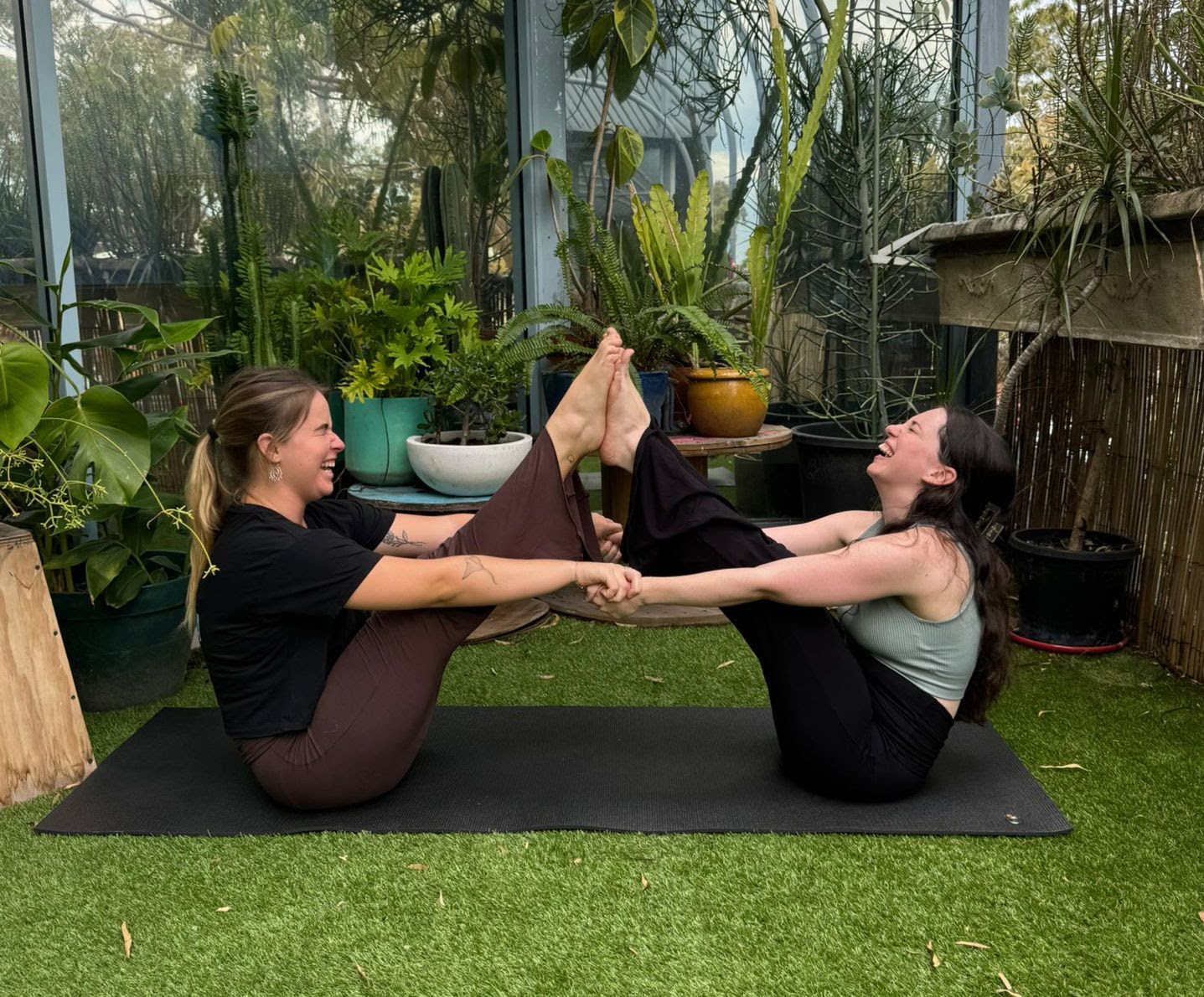 Two women laughing and holding hands as they try and do a yoga pose together.