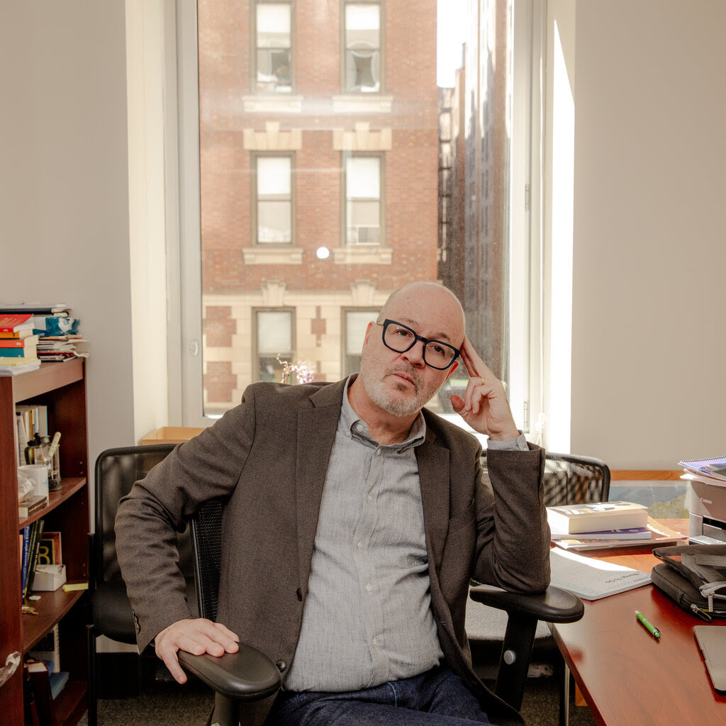 Dominic Preziosi, wearing glasses, sits in an office chair, leaning to his left, his fingers on his temple.