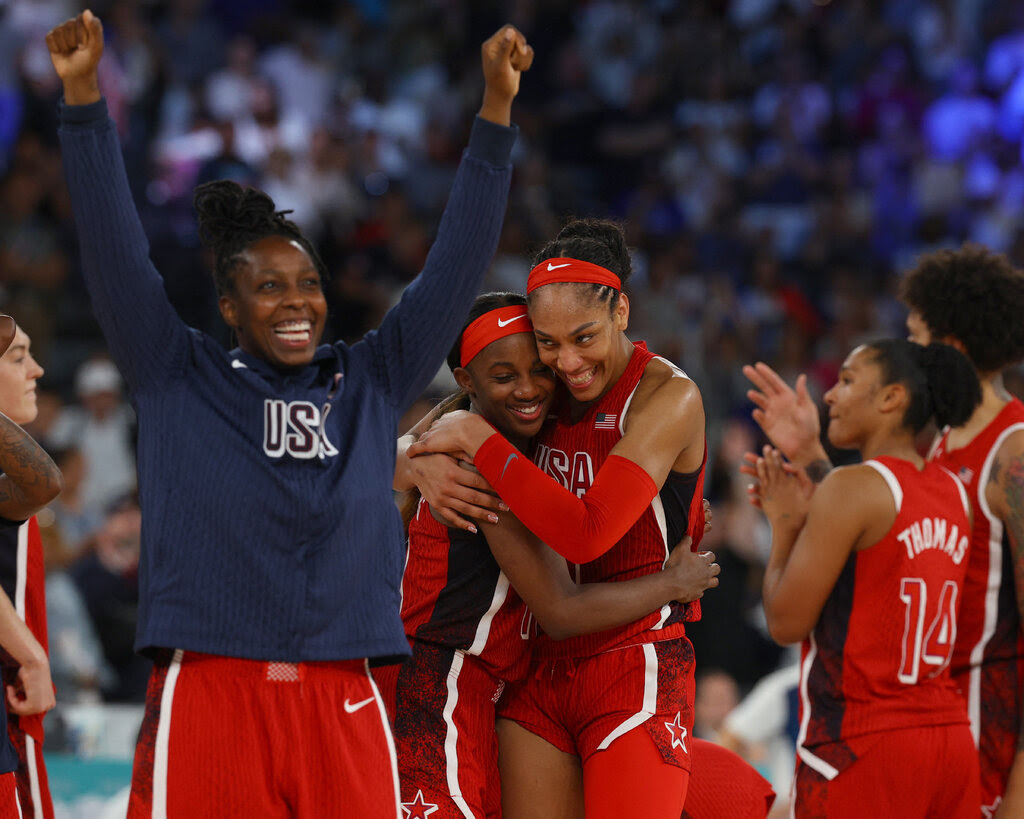 Two members of the United States women’s basketball team in red uniforms embrace, as others behind them clap and one in front of them raises her arms over her head, with a big smile on her face.