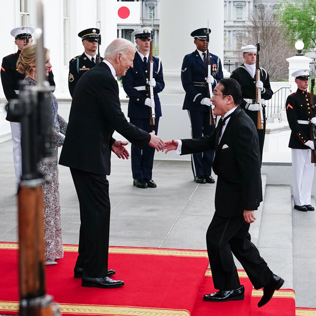 President Biden and the first lady, Dr. Jill Biden, extend hands to Prime Minister Fumio Kishida on a red carpet on the steps of the White House, as six members of an honor guard looked on.