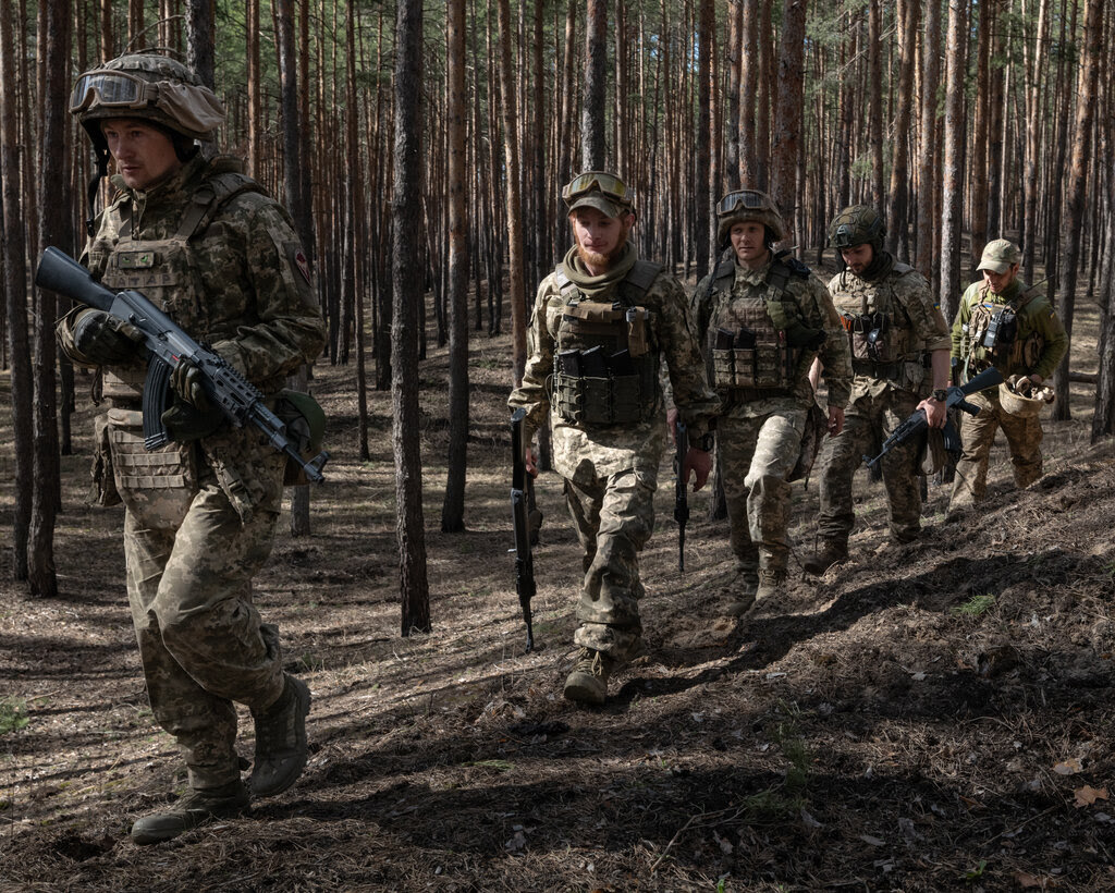 People wearing military uniforms and carrying weapons march in line through a wooded area.