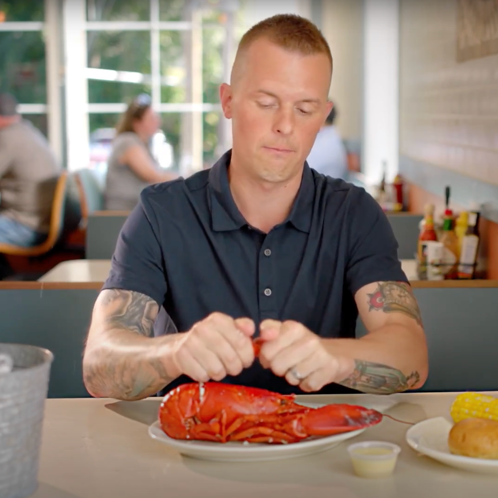 Jared Goldman sitting in a restaurant and looking down as he cracks open a lobster with his hands. He is wearing a short-sleeve shirt that reveals several tattoos on his arms. 