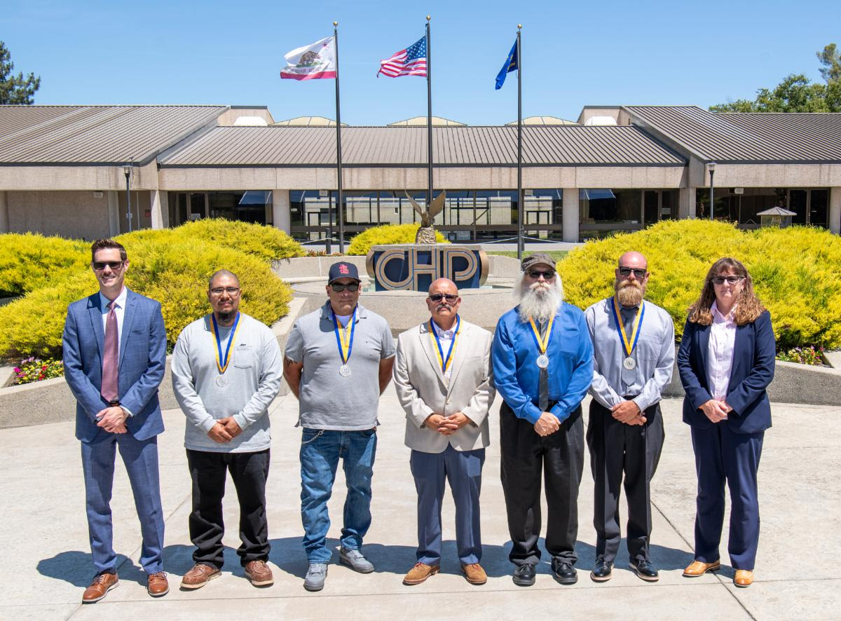Picture shows six men and one woman standing in a line in front of the California Highway Patrol Academy in West Sacramento. Behind them is the CHP Logo with the California and American flags on flag poles. 