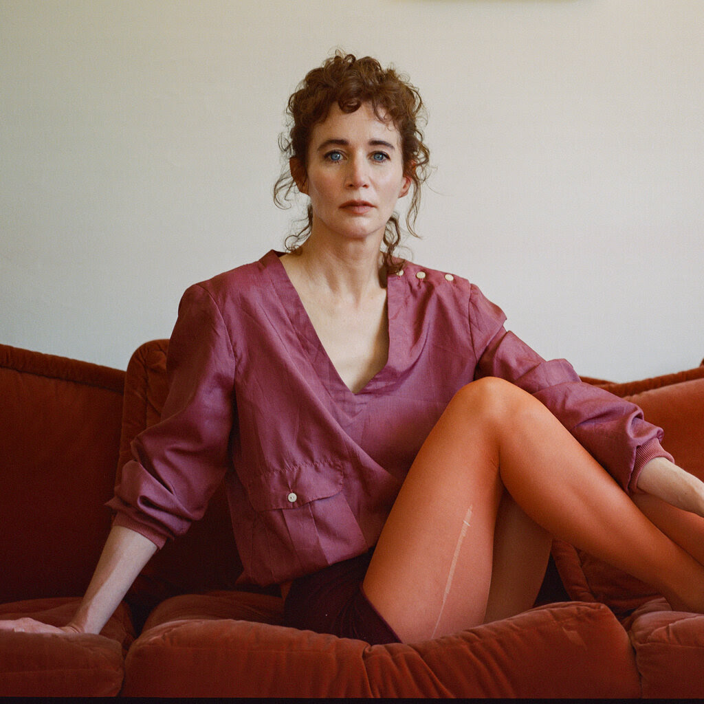 Miranda July seated, posing on an orange velvet couch. She is wearing a mauve top and red tights with a run on the thigh.