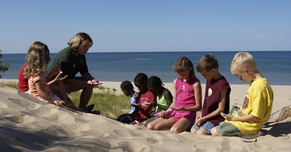 a woman in dark green shirt and khakis explores a sand dune with group of young kids at Hoffmaster State Park. Big blue lake in background