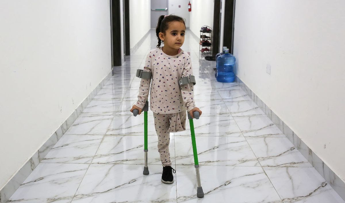 Photo of Gazal Bakr, a Palestinian child from Gaza, with one amputated leg, standing with crutches, in the hallway of the apartment she now lives in. 