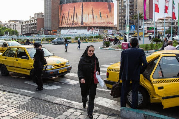 A woman walks past two yellow taxis on a street. There is a billboard in the background with an image of missiles.
