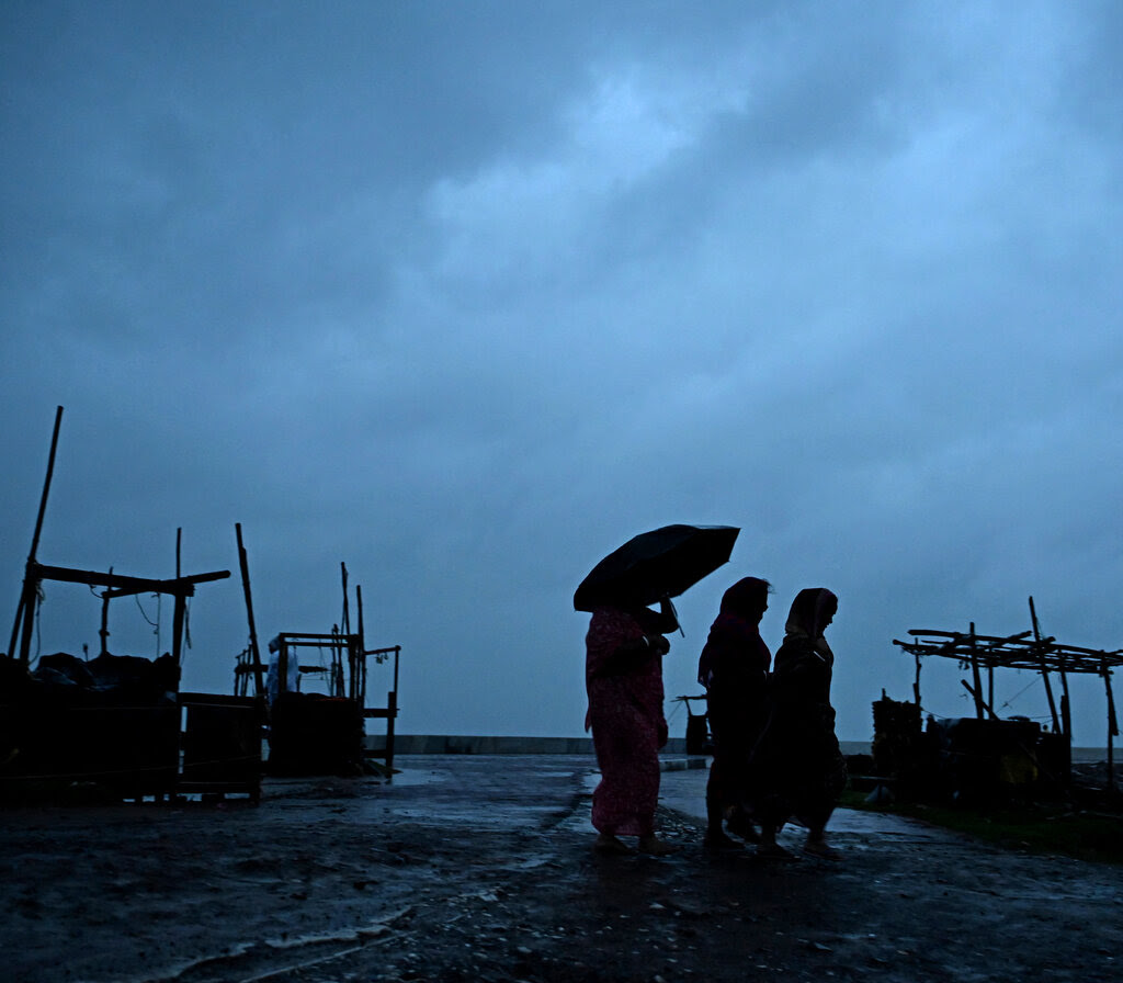 Three silhouetted people, one carrying an umbrella, walk past vendor stalls on a beach with a darkened, cloudy sky in the background.