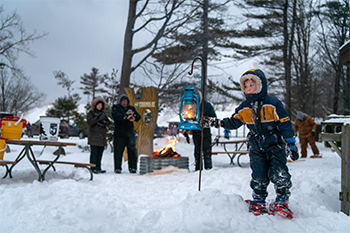 a young boy in blue snowsuit touches a teal lantern on a pole in the snow, with a campfire and a few adults in winter gear in background