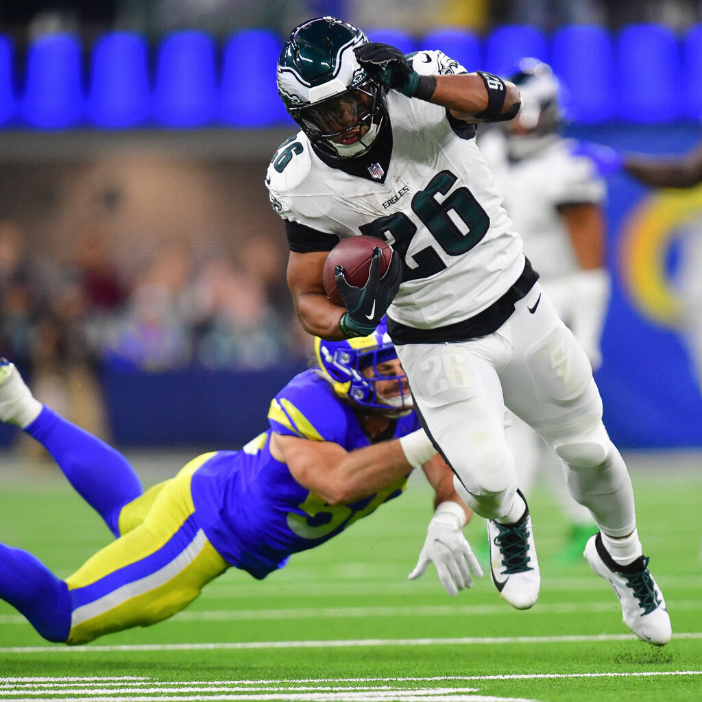 A football player in a No. 26 Eagles uniform holds a football as he runs past a diving player from the Rams.