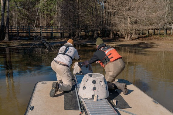 Two men on a flat decked boat push a circular concrete structure from rollers into the water along the shore of Lake Murray