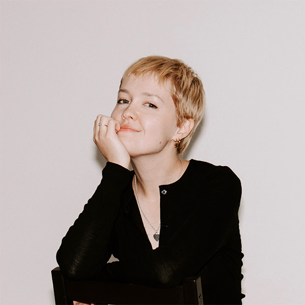 A woman sitting backwards on a chair with her head resting in her hand, smiling at the camera