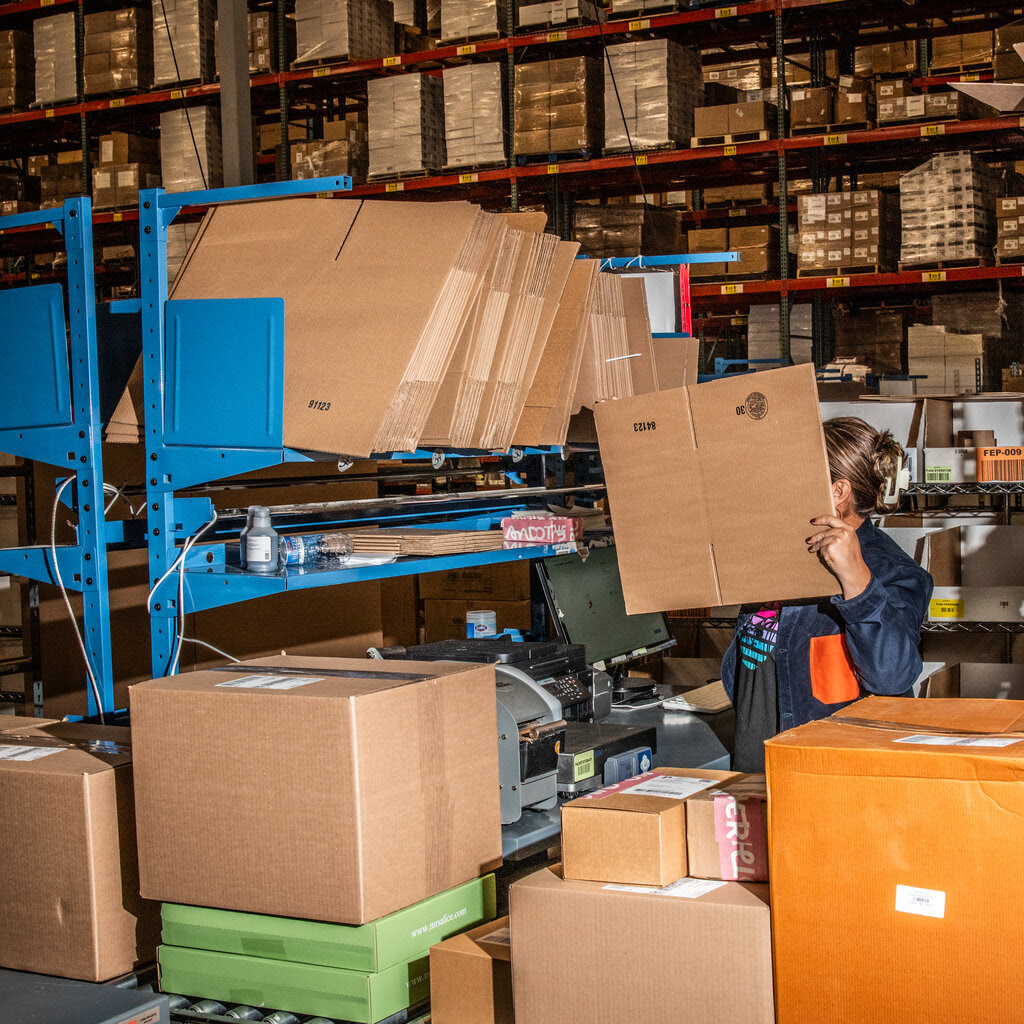 A woman in a warehouse holds up a cardboard box. Her head is only partly visible behind the box.