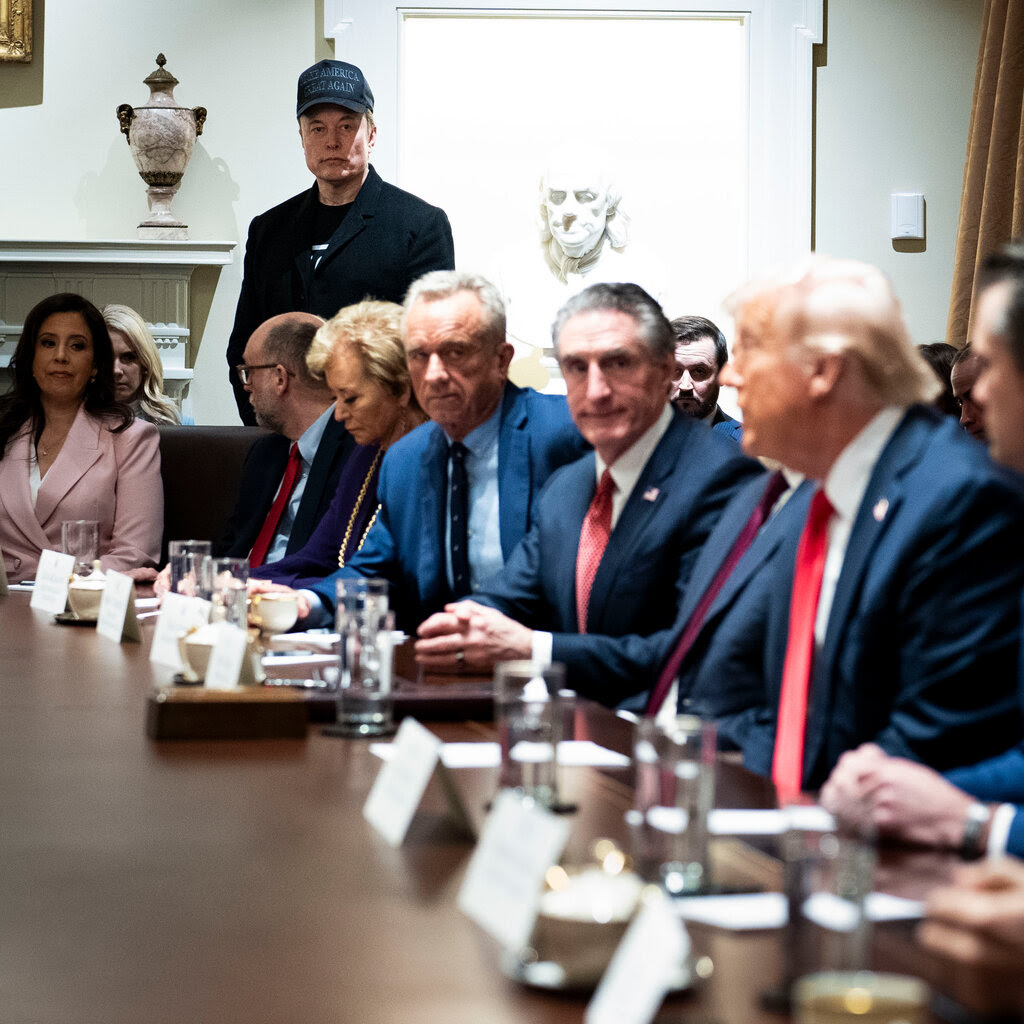 A large table with people sitting next to Donald Trump. Elon Musk standing in the background.