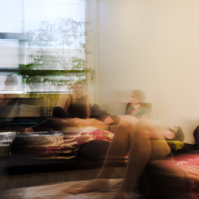 Four or five people sitting or lying on the floor in a white-walled room with a plant near a wide, sunny window. The photograph is distorted in such a way that everything appears blurry, as if the camera had been jerked suddenly to the side just at the moment of exposure. 