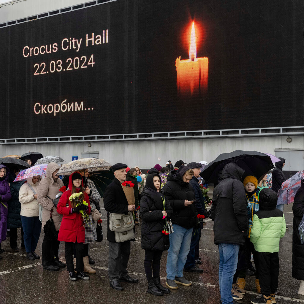 A line of people holding umbrellas and flowers stand before an image of a candle at Crocus City Hall in Moscow.