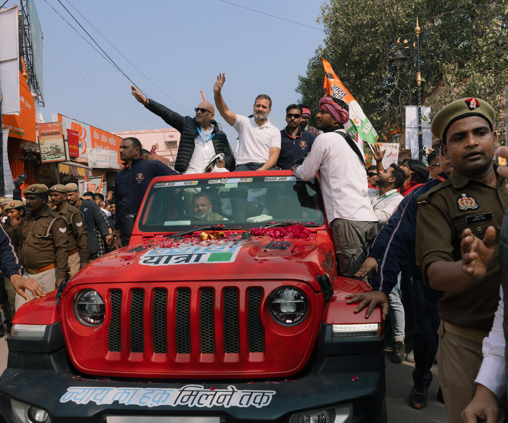 Two men sitting on top of a red Jeep covered in writing wave at crowds.