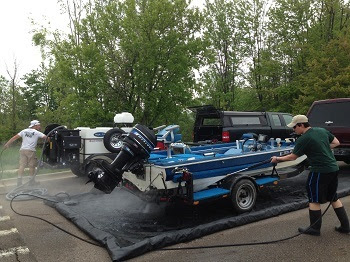 Two men use a portable power washer to clean a boat on a trailer in a parking lot.