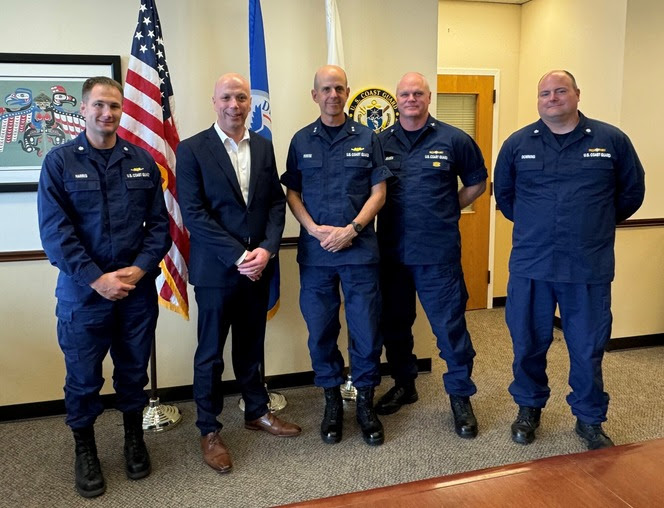 Five people posing for a photo with flags behind and four in Coast Guard uniforms