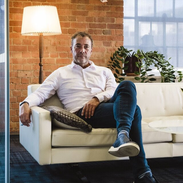 A portrait of Will Lewis, wearing jeans and an open-collar shirt, sitting on a couch with a lamp and brick wall behind him. 